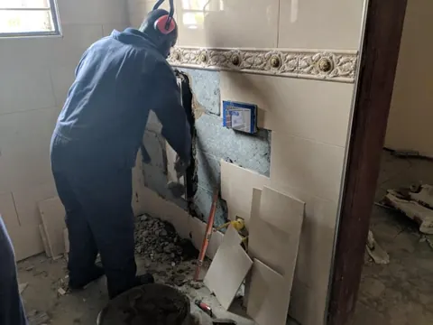 Technician opening a tiled wall with a breaker to access concealed pipework