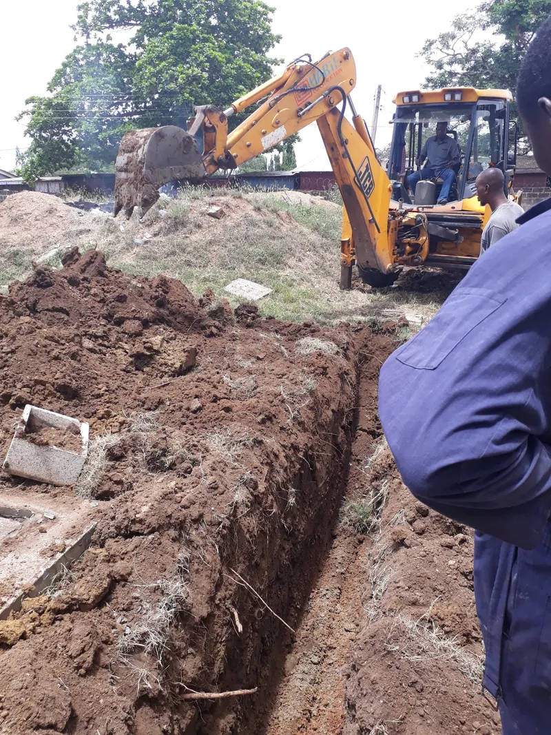 Technician standing in a freshly dug trench beside an excavator, preparing to lay pipe